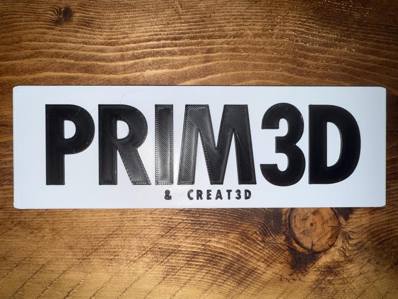 Pu&ograve; includere: Un cartello rettangolare bianco con la scritta "PRIM3D" in grandi lettere nere stampate in 3D. Sotto, il testo pi&ugrave; piccolo recita "& CREATE3D". Il cartello &egrave; posto su uno sfondo di legno.
