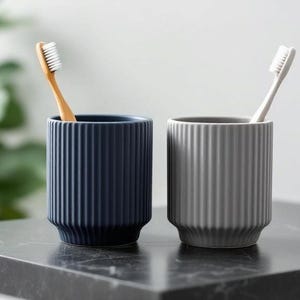 May include: Two ceramic toothbrush holders, one navy blue and one gray, each with vertical ribbed detailing. A wooden toothbrush is in the blue holder, and a white toothbrush is in the gray holder. The holders sit on a dark countertop.