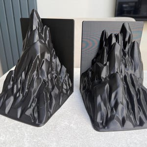 May include: Two black mountain-shaped bookends. Each bookend features a detailed, textured design resembling a rugged mountain range. The bookends are made of a solid material and are designed to stand upright on a flat surface.