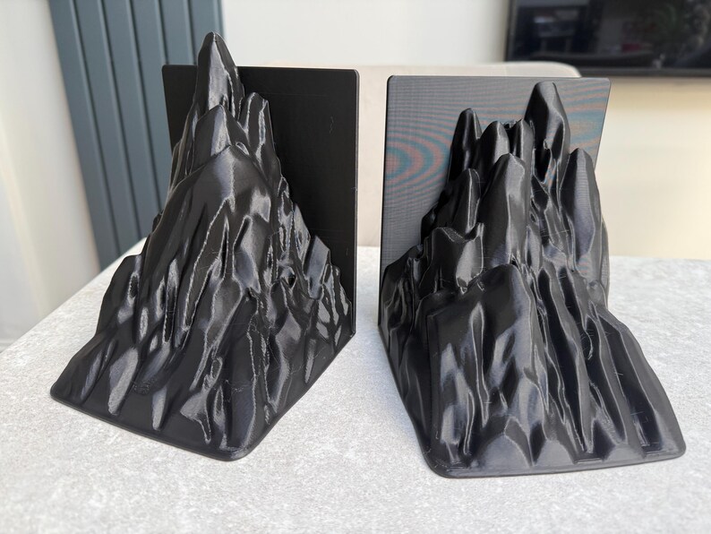 May include: Two black mountain-shaped bookends. Each bookend features a detailed, textured design resembling a rugged mountain range. The bookends are made of a solid material and are designed to stand upright on a flat surface.