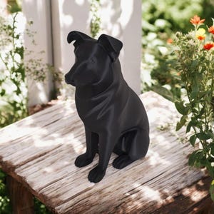 May include: A black dog statue in a sitting position. The sculpture is made of a smooth material and is placed on a weathered wooden surface. The dog has upright ears and a focused gaze. The background includes greenery and a white structure.