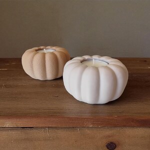 May include: Two pumpkin-shaped candle holders. One is a light peach color, and the other is white. Both have a candle inside. The candle holders are sitting on a wooden surface.