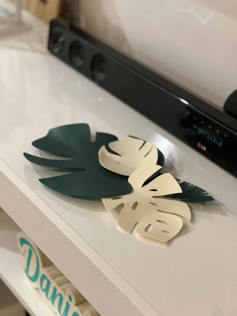May include: Three decorative paper Monstera leaves in dark green and cream colors. The leaves are arranged on a white surface, with a black soundbar in the background. The leaves are cut with detailed edges.