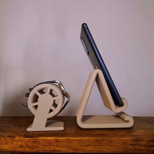 Puede incluir: Dos soportes de tel&eacute;fono de madera. Uno es un soporte triangular que sostiene un tel&eacute;fono negro. El otro es un soporte redondo con un asa de metal plateado. Ambos soportes son de color marr&oacute;n claro y est&aacute;n sobre una superficie de madera.