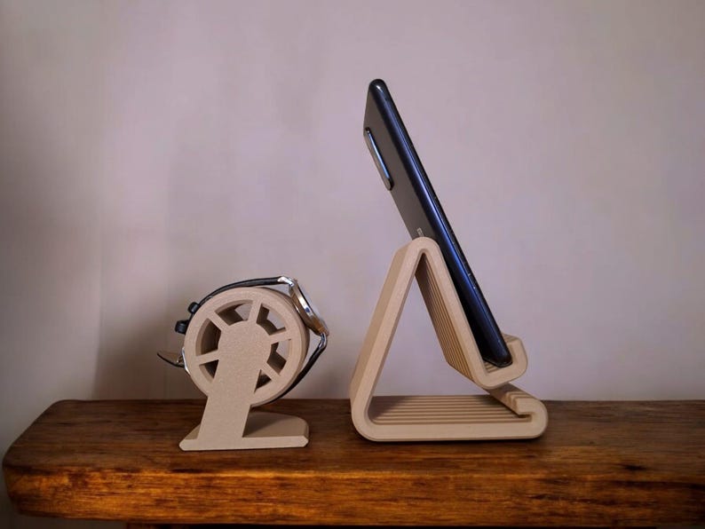 Puede incluir: Dos soportes de tel&eacute;fono de madera. Uno es un soporte triangular que sostiene un tel&eacute;fono negro. El otro es un soporte redondo con un asa de metal plateado. Ambos soportes son de color marr&oacute;n claro y est&aacute;n sobre una superficie de madera.