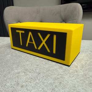 May include: A bright yellow taxi sign with the word "TAXI" in bold, black letters. The sign is rectangular and sits on a gray surface. The background includes a gray chair and a dark gray panel.