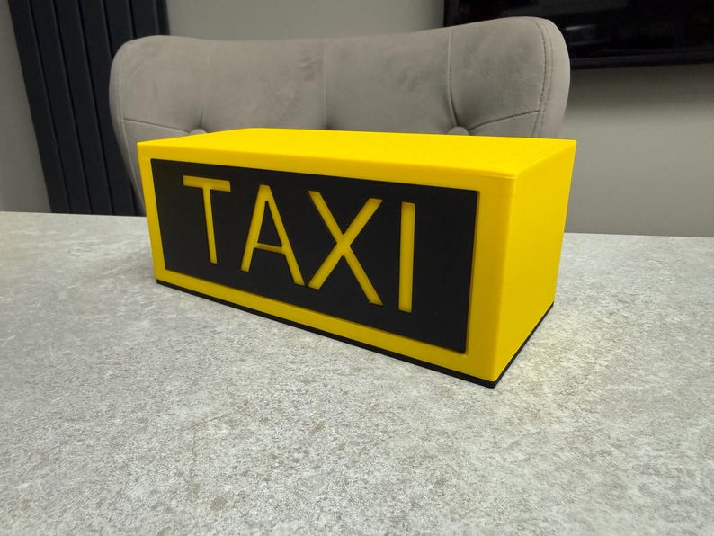 May include: A bright yellow taxi sign with the word "TAXI" in bold, black letters. The sign is rectangular and sits on a gray surface. The background includes a gray chair and a dark gray panel.