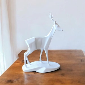 Puede incluir: Una escultura de ciervo geom&eacute;trico blanco. El ciervo est&aacute; de pie sobre una base ovalada blanca, con un cuello delgado y astas. La escultura est&aacute; hecha de un material blanco s&oacute;lido y se coloca sobre una superficie de madera.