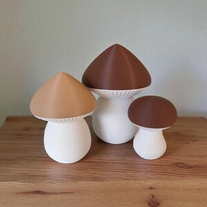 May include: Three decorative mushroom figurines in varying sizes. The caps are brown and tan, while the stems are white. The mushrooms are arranged on a wooden surface, creating a whimsical display.