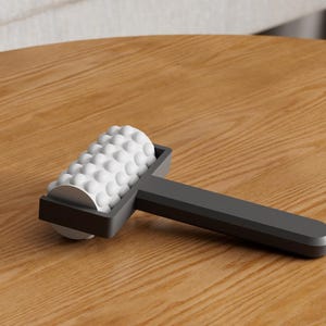 May include: A gray massage roller with a white, textured surface. The roller is designed with a series of small, rounded bumps. The handle is a solid gray color. The item is resting on a wooden surface.