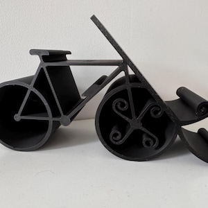 May include: A black bicycle-shaped wine bottle holder. The holder features a stylized design with circular wheels and a seat. The wheels have decorative scrollwork. The holder is made of a solid, matte black material.