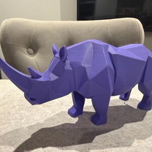 May include: A geometric, purple rhinoceros sculpture. The rhino is a solid color with a faceted design, giving it a modern, angular appearance. The sculpture has a prominent horn and is positioned on a light-colored surface.