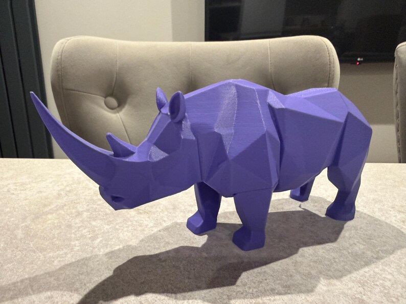 May include: A geometric, purple rhinoceros sculpture. The rhino is a solid color with a faceted design, giving it a modern, angular appearance. The sculpture has a prominent horn and is positioned on a light-colored surface.