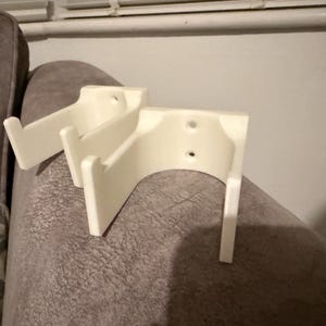 Op de afbeelding: Twee off-white, 3D-geprinte wandbevestigingen. Elke bevestiging heeft een gebogen achterkant met twee gaten en twee verticale steunen. De bevestigingen zijn ontworpen om een ​​item vast te houden, mogelijk een plank of een decoratief stuk. De bevestigingen liggen op een grijze ondergrond.