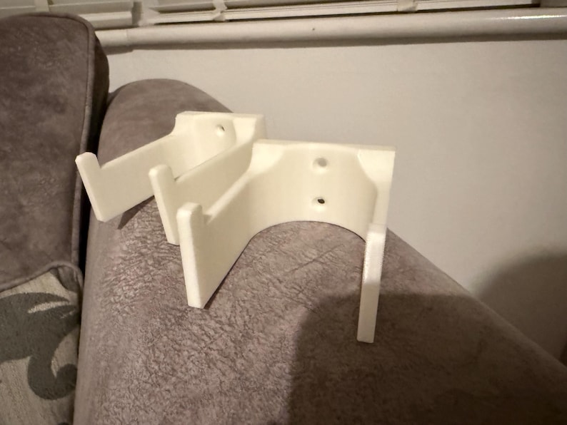 Op de afbeelding: Twee off-white, 3D-geprinte wandbevestigingen. Elke bevestiging heeft een gebogen achterkant met twee gaten en twee verticale steunen. De bevestigingen zijn ontworpen om een ​​item vast te houden, mogelijk een plank of een decoratief stuk. De bevestigingen liggen op een grijze ondergrond.