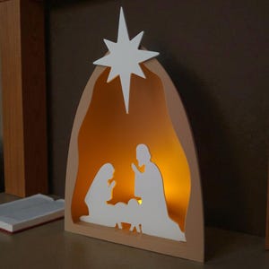 May include: A wooden nativity scene with a white star. The scene features white silhouettes of Mary, Joseph, and baby Jesus, with a warm light inside. The frame is a light tan colour.