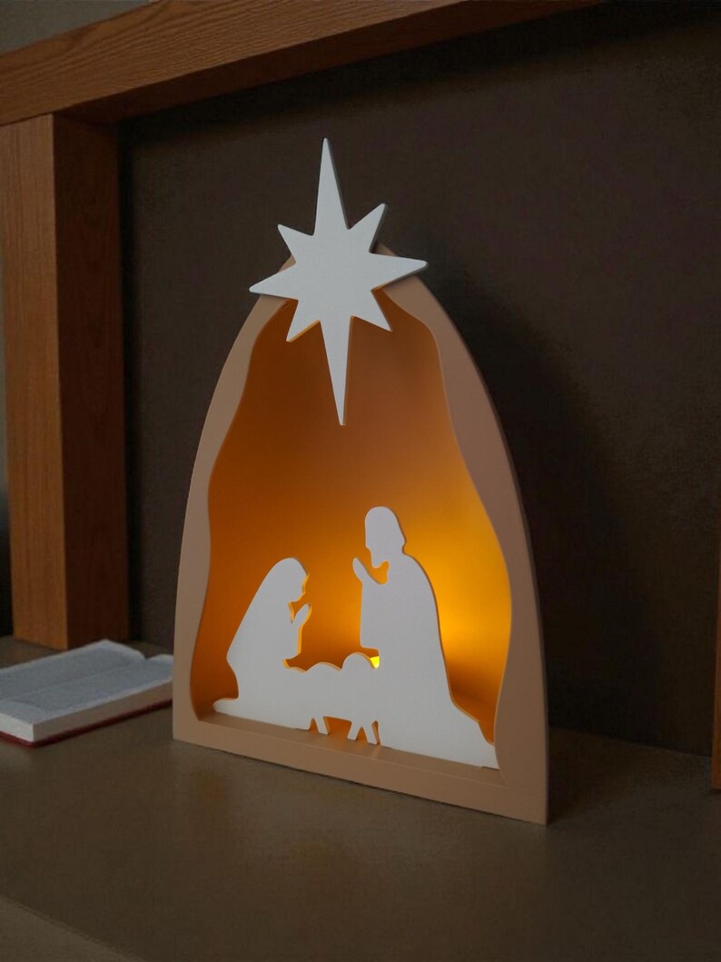 May include: A wooden nativity scene with a white star. The scene features white silhouettes of Mary, Joseph, and baby Jesus, with a warm light inside. The frame is a light tan colour.