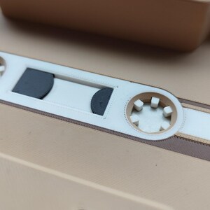 Pu&ograve; includere: Primo piano di una cassetta beige con dettagli bianchi e marroni. Il nastro presenta un design dettagliato con una finestra centrale ed elementi circolari. L'estetica generale &egrave; retr&ograve; e nostalgica.