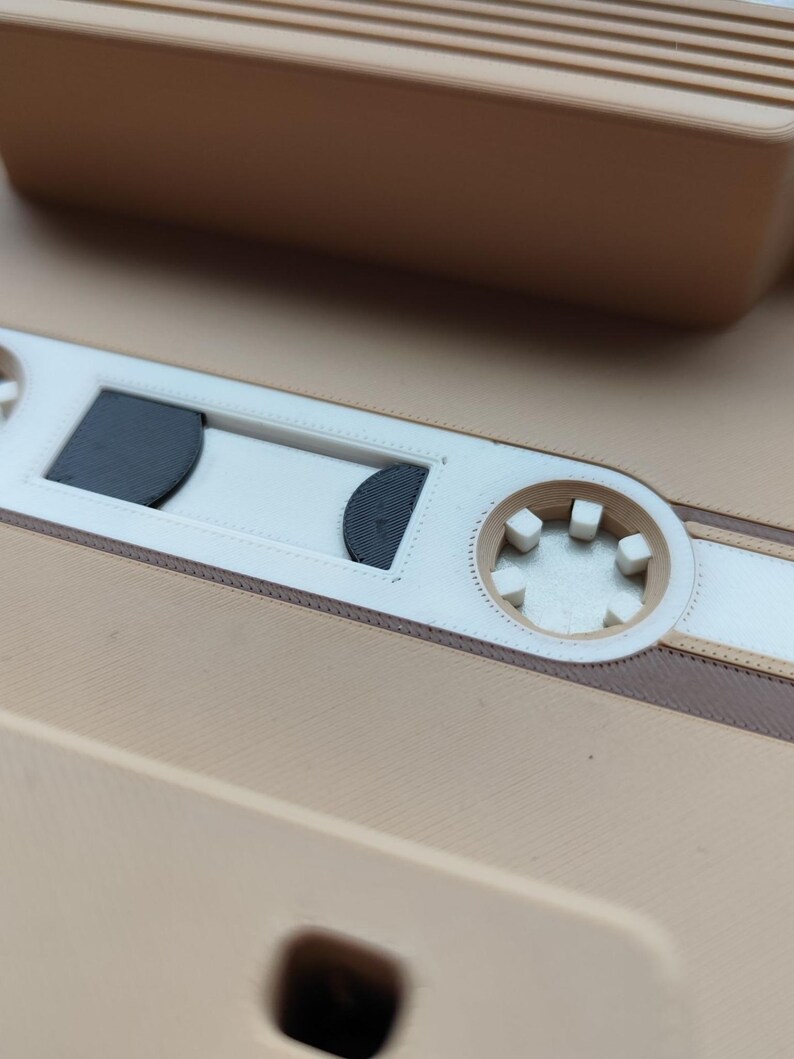 Pu&ograve; includere: Primo piano di una cassetta beige con dettagli bianchi e marroni. Il nastro presenta un design dettagliato con una finestra centrale ed elementi circolari. L'estetica generale &egrave; retr&ograve; e nostalgica.