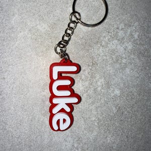 May include: A red and white keychain with the name "Luke" in a bold, rounded font. The keychain has a silver metal ring and chain. The background is a mottled gray surface.