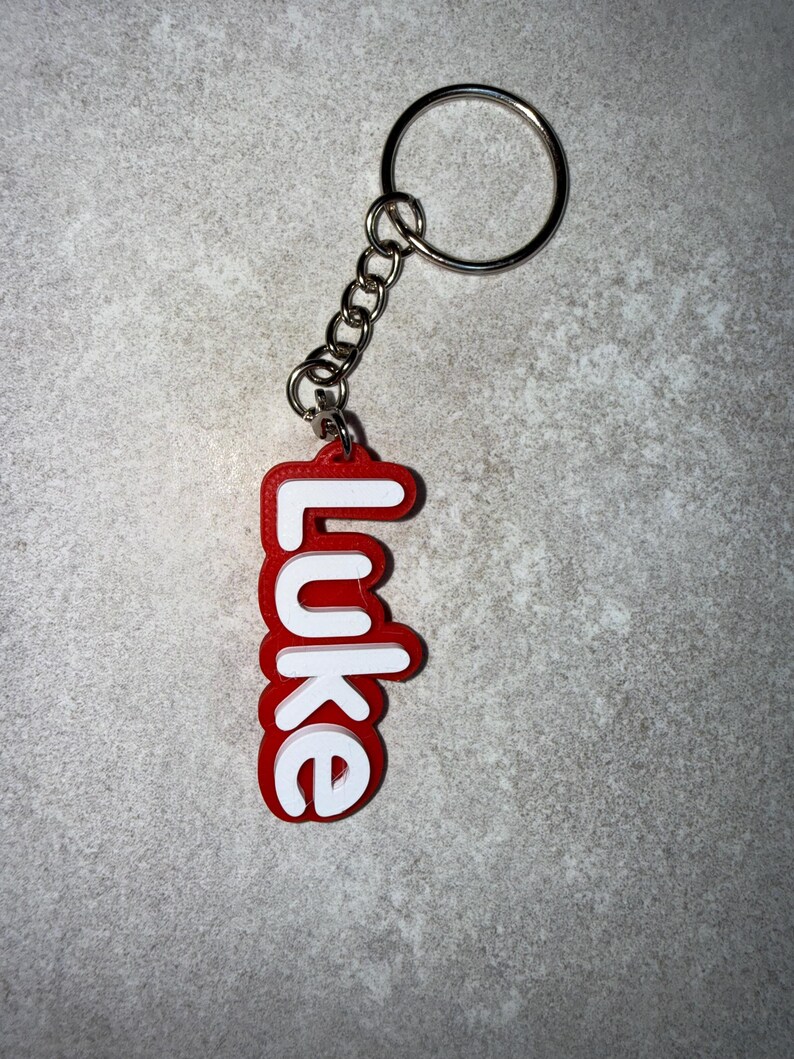 May include: A red and white keychain with the name "Luke" in a bold, rounded font. The keychain has a silver metal ring and chain. The background is a mottled gray surface.