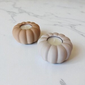 May include: Two pumpkin-shaped candle holders in neutral tones. One is a light beige, the other a muted taupe. Each holds a small white candle. The holders have a ribbed design, creating a textured surface. Perfect for fall decor or adding a cozy touch.