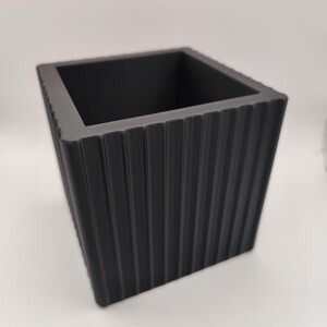 May include: A black square planter with vertical ribbed detailing. The planter has a modern design and a matte finish. The open top allows for planting flowers, herbs, or small plants. The planter is suitable for indoor or outdoor use.