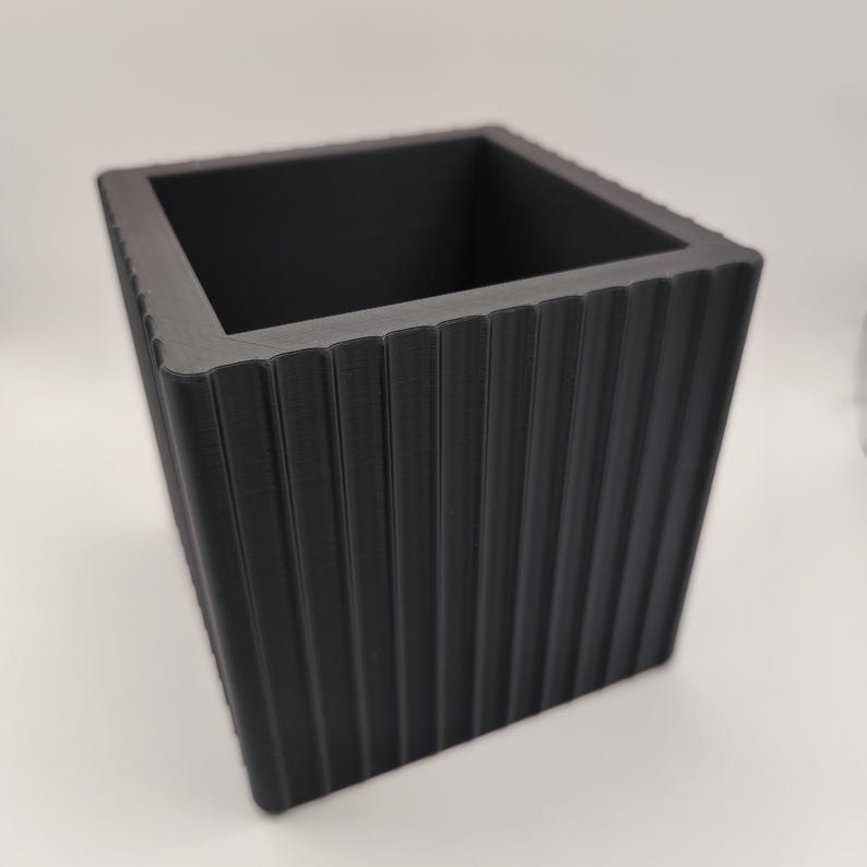 May include: A black square planter with vertical ribbed detailing. The planter has a modern design and a matte finish. The open top allows for planting flowers, herbs, or small plants. The planter is suitable for indoor or outdoor use.