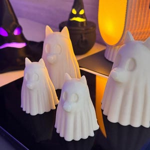 Pu&ograve; includere: Una collezione di figurine bianche a forma di cane, ognuna avvolta in un sudario spettrale e fluente. Le figurine variano in dimensioni e sono disposte su una superficie nera riflettente. Sullo sfondo, un cappello da strega nero e oggetti a forma di calderone.