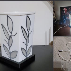 May include: A white rectangular lamp with a black base features a leaf design in grey and black. The lamp is illuminated, showcasing the leaf pattern. The lamp is connected to a power source via a white cord. Home decor.