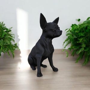 May include: A black Chihuahua statue sits on a light wood surface, with green plants in the background. The dog figurine has large ears and is posed in a seated position, facing to the right.