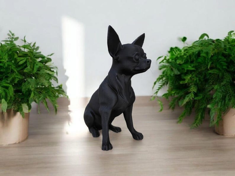 May include: A black Chihuahua statue sits on a light wood surface, with green plants in the background. The dog figurine has large ears and is posed in a seated position, facing to the right.