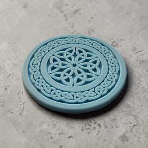 May include: A light blue, round coaster with a Celtic knot design. The coaster features a central knot pattern surrounded by a circular border of interwoven knots. The coaster is approximately 4 inches in diameter.