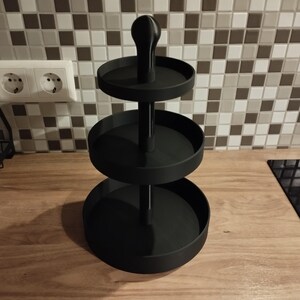 May include: A black, three-tiered serving tray with a central handle. The tray has three circular platforms of decreasing size, supported by a central pole. The handle is rounded. The tray is made of a matte black material.