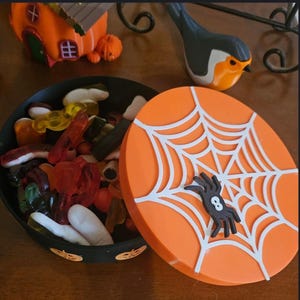 May include: An orange Halloween candy dish with a white spiderweb design and a black spider with the number 8. The dish is filled with colourful gummy sweets. A small orange house and bird figurine are in the background.