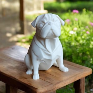 K&ouml;nnte beinhalten: Eine wei&szlig;e Mops-Hundestatue mit geometrischem Design, die auf einem Holztisch sitzt. Die Statue hat ein detailliertes Gesicht und ist vor einem verschwommenen Hintergrund aus Gr&uuml;n und Blumen platziert.
