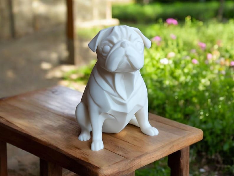 K&ouml;nnte beinhalten: Eine wei&szlig;e Mops-Hundestatue mit geometrischem Design, die auf einem Holztisch sitzt. Die Statue hat ein detailliertes Gesicht und ist vor einem verschwommenen Hintergrund aus Gr&uuml;n und Blumen platziert.