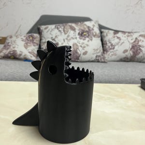 May include: A black, dinosaur-shaped container with a wide open mouth and a fin. The container has a cylindrical body with a jagged edge around the top, resembling teeth. The container is sitting on a light-colored surface.