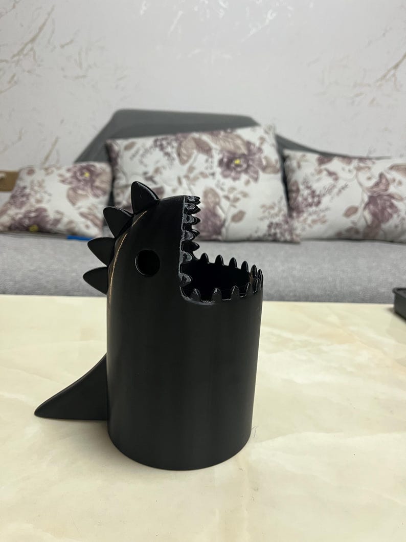 May include: A black, dinosaur-shaped container with a wide open mouth and a fin. The container has a cylindrical body with a jagged edge around the top, resembling teeth. The container is sitting on a light-colored surface.