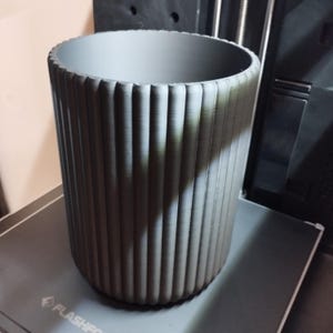 May include: A cylindrical, gray planter with vertical ribbed detailing. The planter has a wide opening and a textured surface. The design is modern and minimalist, suitable for indoor or outdoor use. The planter is likely made of a durable material.