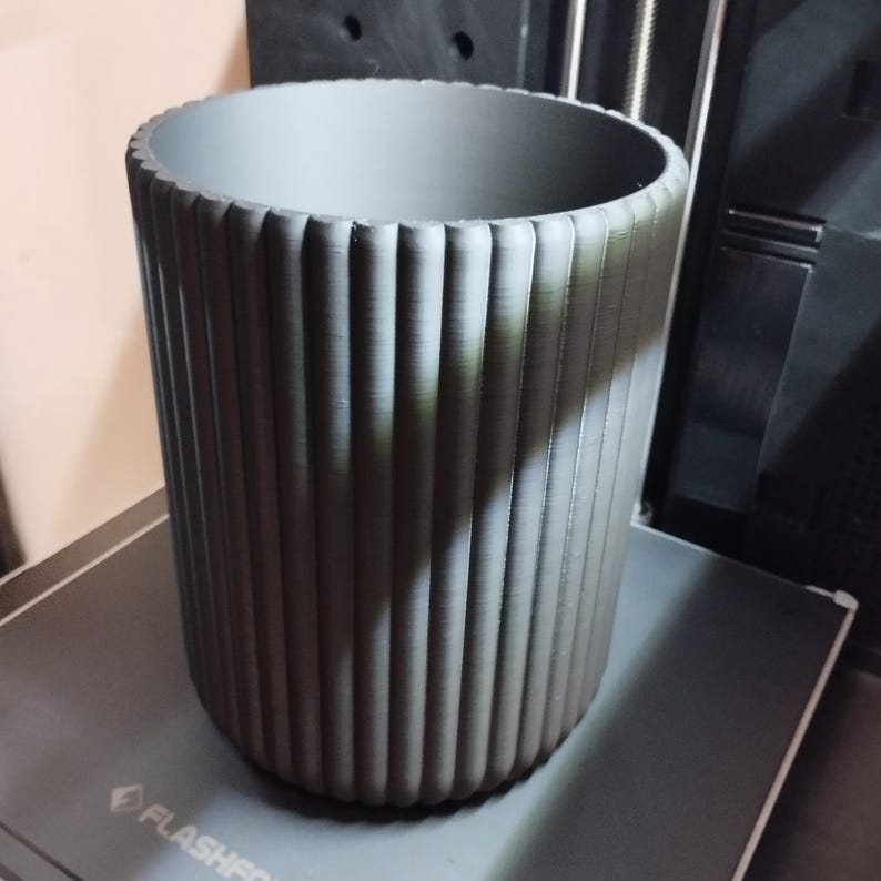 May include: A cylindrical, gray planter with vertical ribbed detailing. The planter has a wide opening and a textured surface. The design is modern and minimalist, suitable for indoor or outdoor use. The planter is likely made of a durable material.