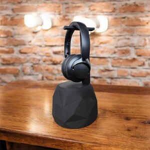May include: Black over-ear headphones resting on a black geometric headphone stand. The stand is on a wooden table, with a brick wall and lights in the background. The headphones appear to be made of plastic.