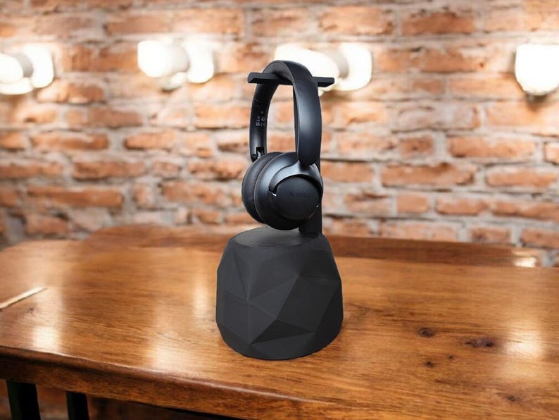 May include: Black over-ear headphones resting on a black geometric headphone stand. The stand is on a wooden table, with a brick wall and lights in the background. The headphones appear to be made of plastic.