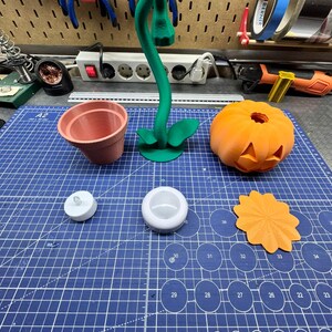 May include: A collection of 3D-printed items on a blue cutting mat. Includes a terracotta-colored flower pot, a green plant, a carved orange pumpkin, a white LED candle, a white candle holder, and an orange decorative piece. Tools are visible in the background.