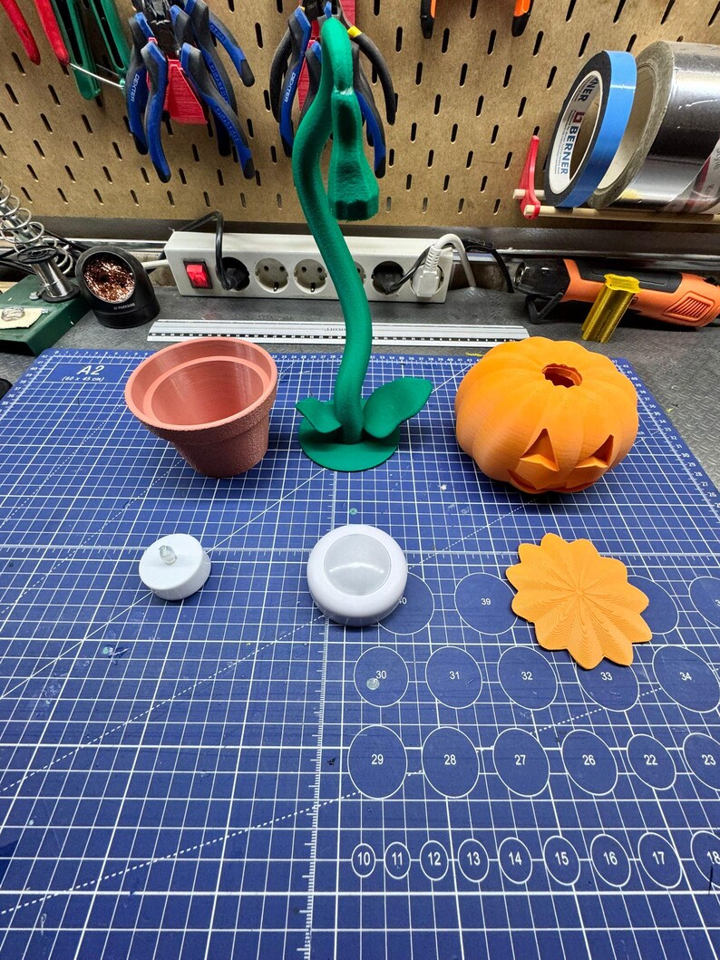 May include: A collection of 3D-printed items on a blue cutting mat. Includes a terracotta-colored flower pot, a green plant, a carved orange pumpkin, a white LED candle, a white candle holder, and an orange decorative piece. Tools are visible in the background.