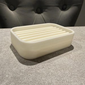 May include: A rectangular, off-white soap dish with a slatted top. The dish has rounded corners and sits on a gray surface. The soap dish is a bathroom accessory.