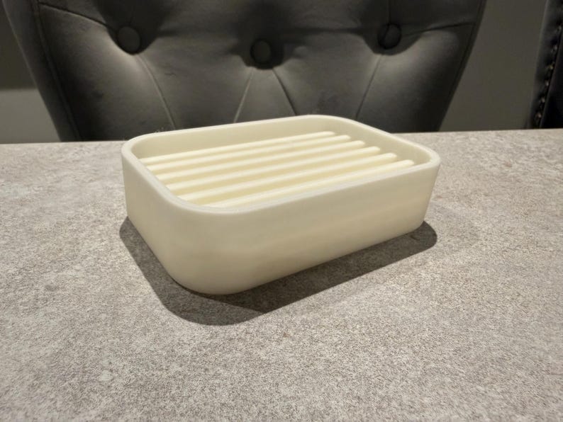 May include: A rectangular, off-white soap dish with a slatted top. The dish has rounded corners and sits on a gray surface. The soap dish is a bathroom accessory.