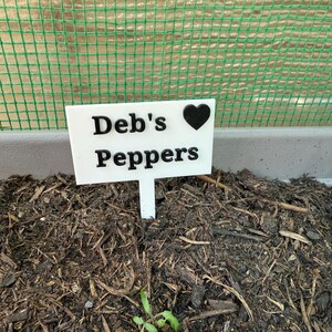 Puede incluir: Un marcador de plantas blanco con el texto "Deb's Peppers" y un s&iacute;mbolo de coraz&oacute;n negro. El marcador se coloca en tierra oscura con una peque&ntilde;a planta verde brotando. Una valla de malla verde est&aacute; en el fondo.