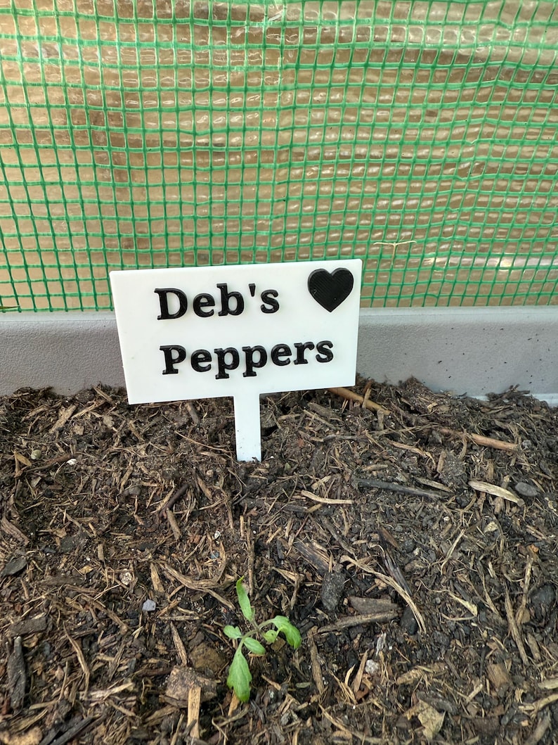 Puede incluir: Un marcador de plantas blanco con el texto "Deb's Peppers" y un s&iacute;mbolo de coraz&oacute;n negro. El marcador se coloca en tierra oscura con una peque&ntilde;a planta verde brotando. Una valla de malla verde est&aacute; en el fondo.