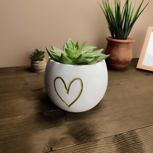 Op de afbeelding: Een witte, ronde keramische plantenbak met een gouden hartontwerp, met een groene vetplant. Andere planten in terracotta potten zijn zichtbaar, wat een natuurlijk, decoratief thema cre&euml;ert.